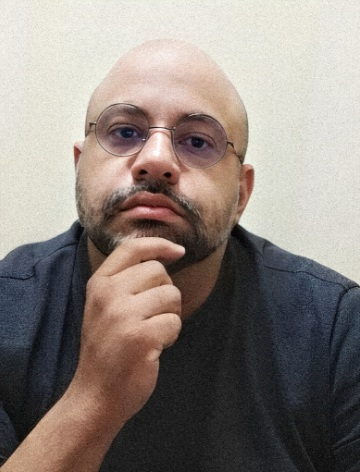 Na imagem, o terapeuta online Herlon Vasconcelos aparece com expressão pensativa, apoiando o queixo na mão. Ele é um homem branco, careca, com barba grisalha, óculos redondos e veste uma camisa cinza escura. Parece ter cerca de 30 anos.